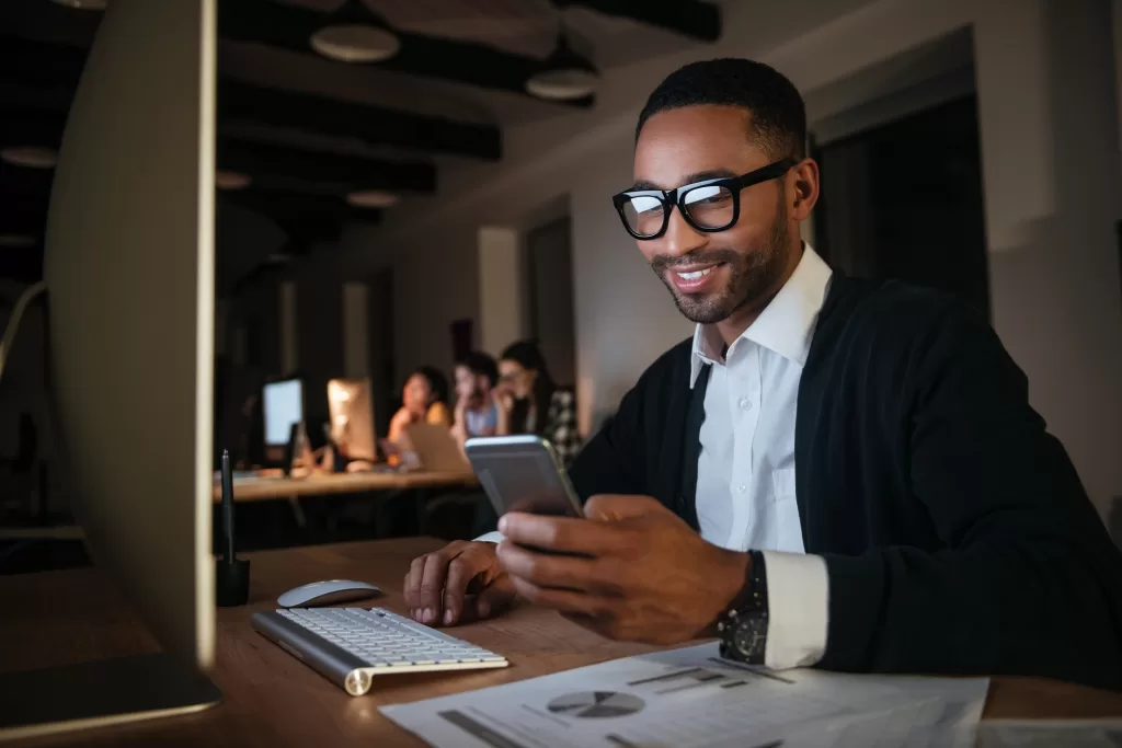 Man looking at texts on his phone while sitting in front of his computer in a darkened room.
